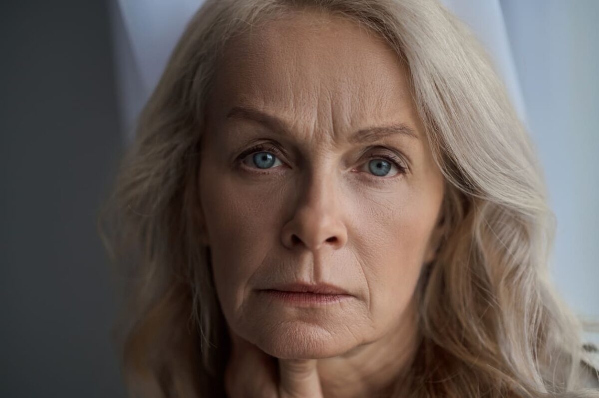 Close-up of a middle-aged woman with blue eyes and gray hair, looking contemplative with a hand resting near her neck.
